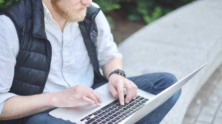 white man with beard sitting on curb typing on computer