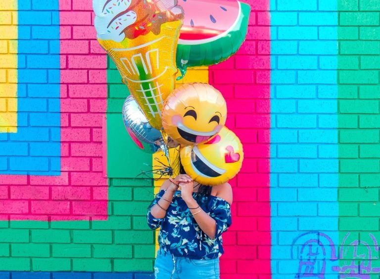 A woman holds multiple emoji balloons in front of a brightly colored wall.