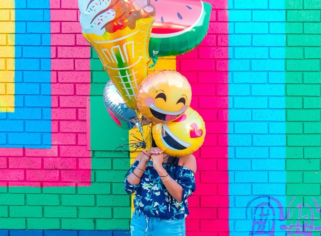 A woman holds multiple emoji balloons in front of a brightly colored wall.